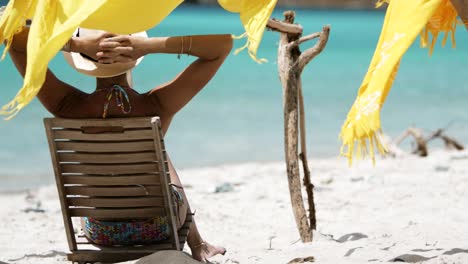 mujer con los brazos detrás de la cabeza sentada en una silla de teca leyendo un libro en una playa tropical