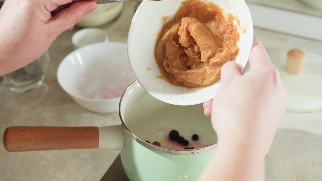 Cook pours mashed ingredient into light green pot with wooden handle containing mixed berries on kitchen counter, preparing homemade dish with natural ingredients