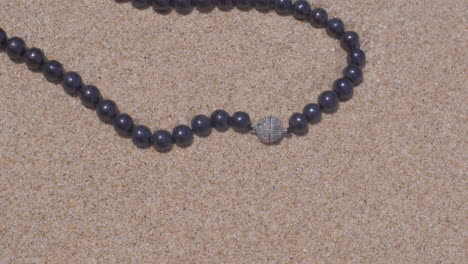 cinematic orbiting shot of a natural pearl necklace with silver jewellery laying on a sand beach close to the ocean, 120 fps, slomo