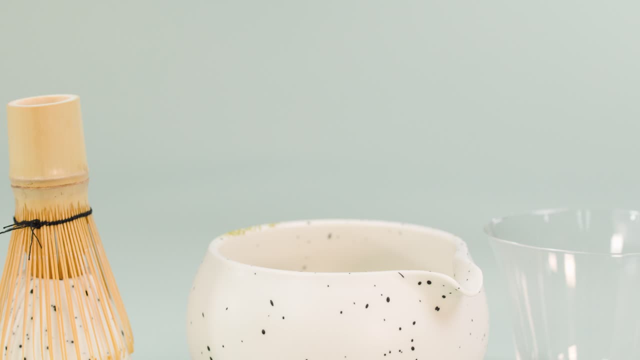 Clear water pours into a speckled ceramic bowl beside bamboo whisk, minimal clean background