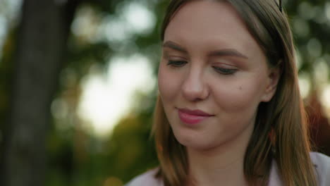 close-up of young lady with glasses resting on her head, looking up contemplatively with warm smile, blurred background features trees with light filtering through them