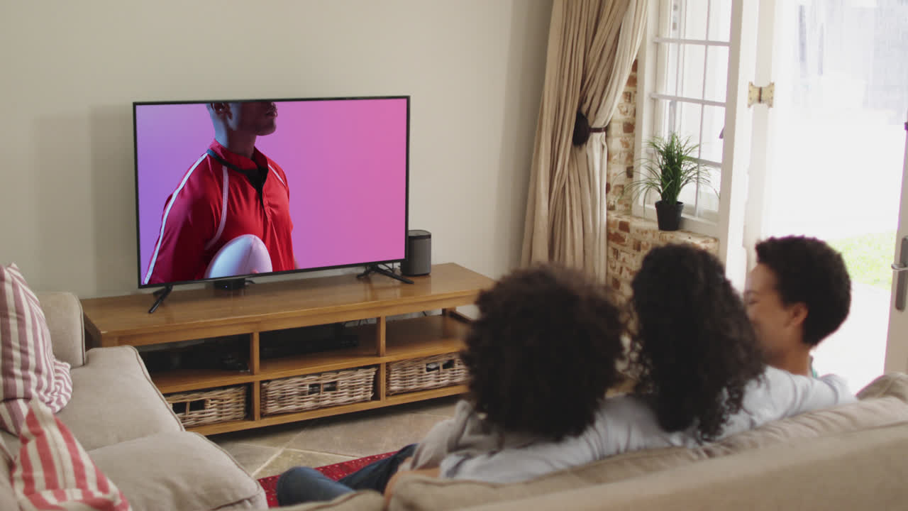 familia biracial viendo la televisión con un jugador de rugby afroamericano con la pelota en la pantalla