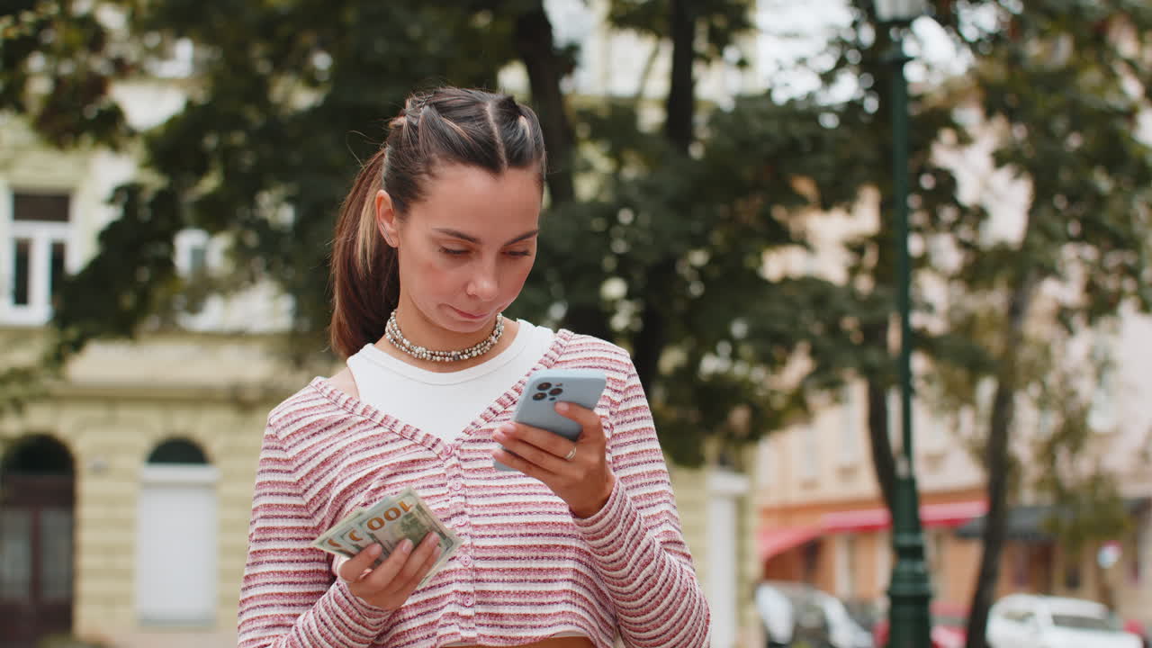 Donna felice che conta soldi, dollari in contanti, usa un'app di calcolatrice per smartphone in una strada urbana.