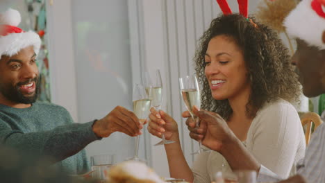 Familia-Multigeneracional-Con-Gorros-De-Papá-Noel-Haciendo-Un-Brindis-Durante-La-Comida-Navideña-En-Casa-Juntos