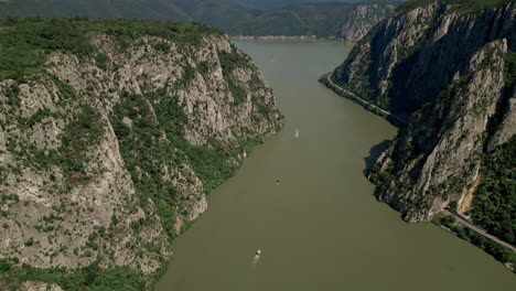 Luftaufnahme-Der-Atemberaubenden-Dubova-Schluchten-In-Rumänien-Mit-Hoch-Aufragenden-Klippen,-Die-Einen-Ruhigen-Fluss-Flankieren,-Mit-Gleitenden-Booten-Und-Einer-Entfernten-Siedlung