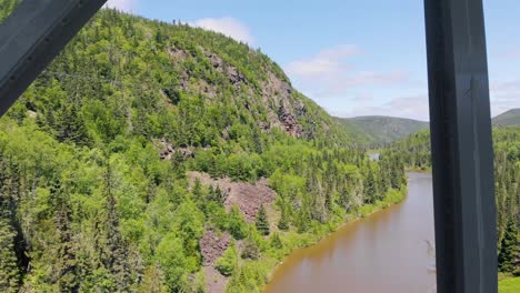 a drone flies slowly through the metal beams of a large bridge high above the water of a river surrounded by trees and mountains on all sides