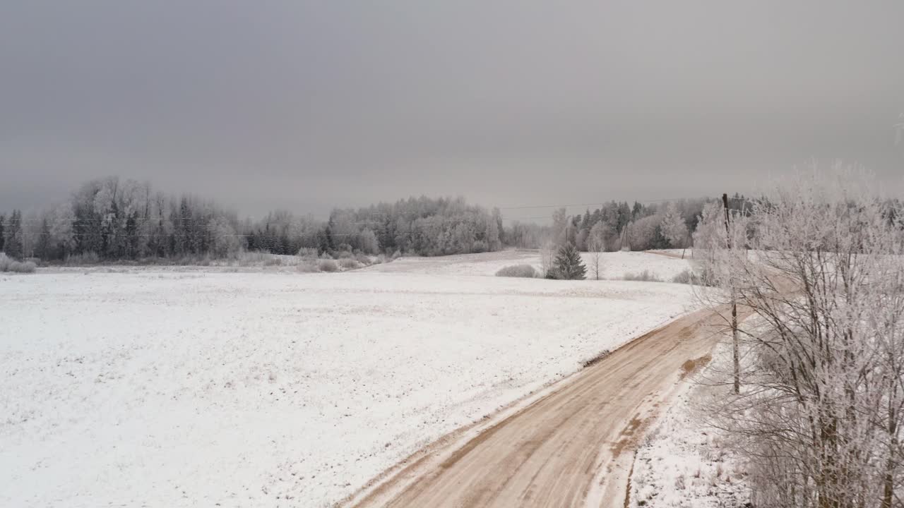Aerial drone shot over gravel road in countryside. Country dirt road in snowy winter with frozen forest in the background.