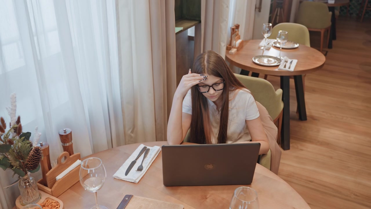 Donna bianca che studia al laptop in un caffè, prendendo appunti e digitando con intenzione, luce solare che filtra attraverso le tende, decorazioni con piante e tavolo organizzato, tazza di caffè e libro di testo nelle vicinanze, in un bistrò calmo.