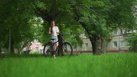 una mujer joven se acerca a su bicicleta estacionada en un camino pavimentado, sosteniendo guantes negros mientras se prepara para ponérselos, el fondo presenta vegetación exuberante, árboles y alguien caminando en la distancia