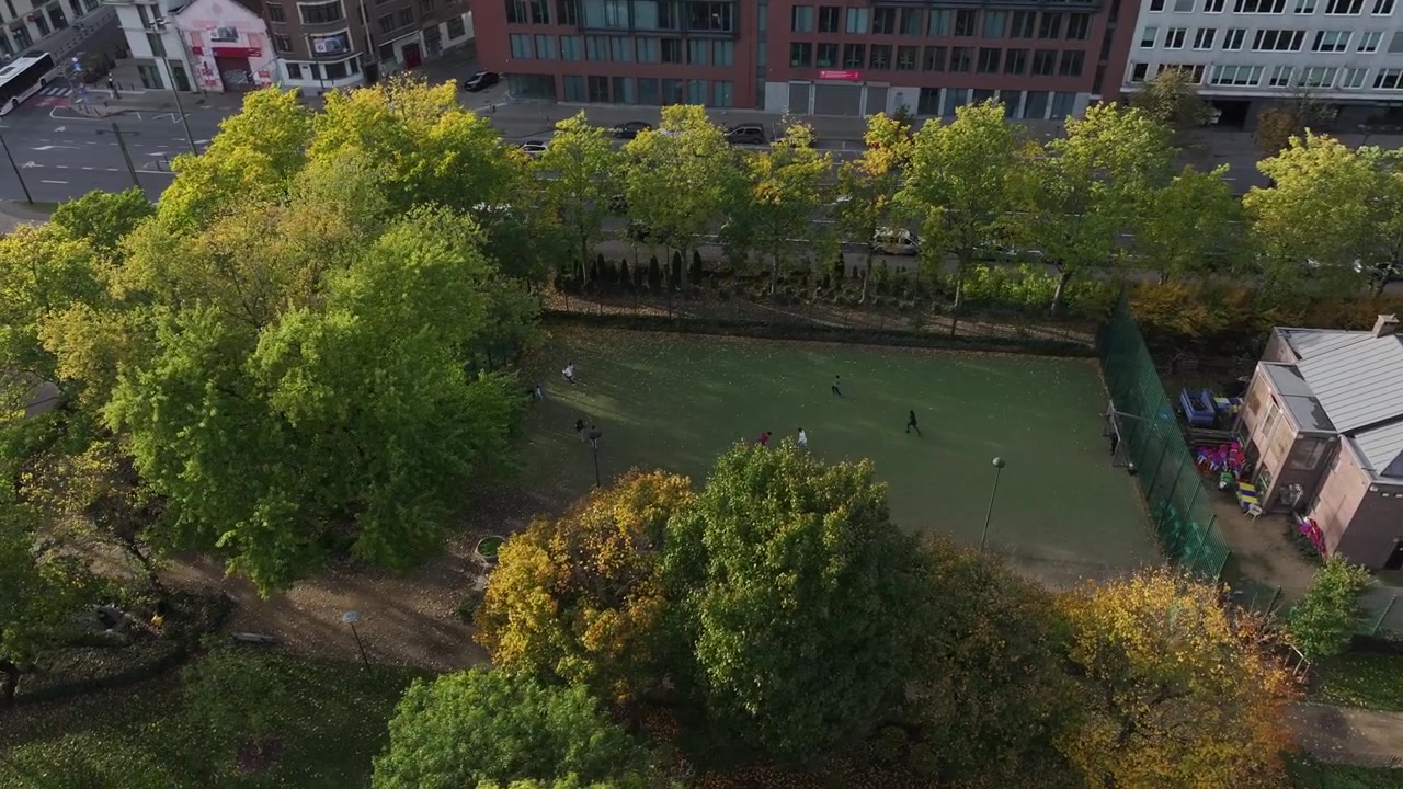 Dronebeelden van een stadspark en voetbalveld waar jongeren spelen, met stedelijke gebouwen op de achtergrond op een zonnige dag.
