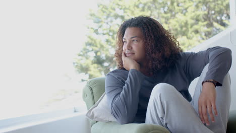 feliz hombre biracial con cabello largo sentado en una habitación soleada disfrutando de la vista, espacio de copia, cámara lenta