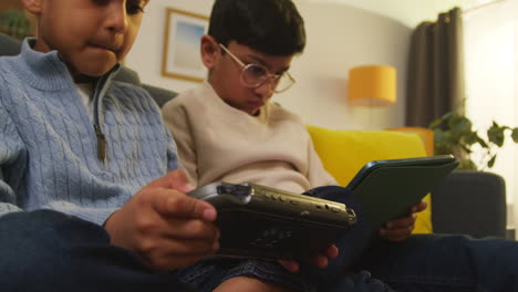 Two-Young-Boys-Sitting-On-Sofa-At-Home-Playing-Games-Or-Streaming-Onto-Digital-Tablet-And-Handheld-Gaming-Device-1