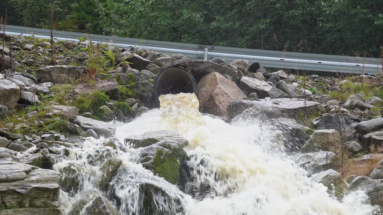 언덕의 도로 울타리 아래의 플라스틱 배수 파이프에서 흘러나오는 갈색 홍수 물 - 폭우 동안 홍수 개념