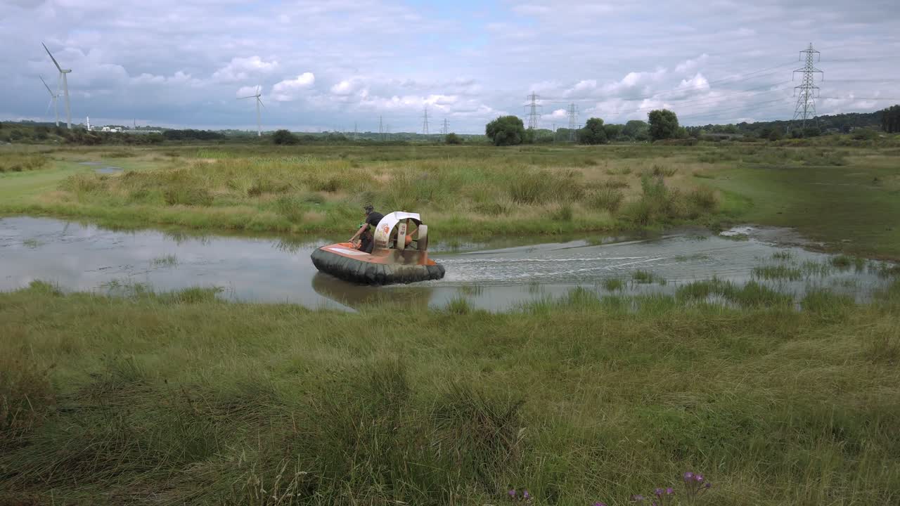 Hovercraft passing through shot over water and grassland with wind turbine in background