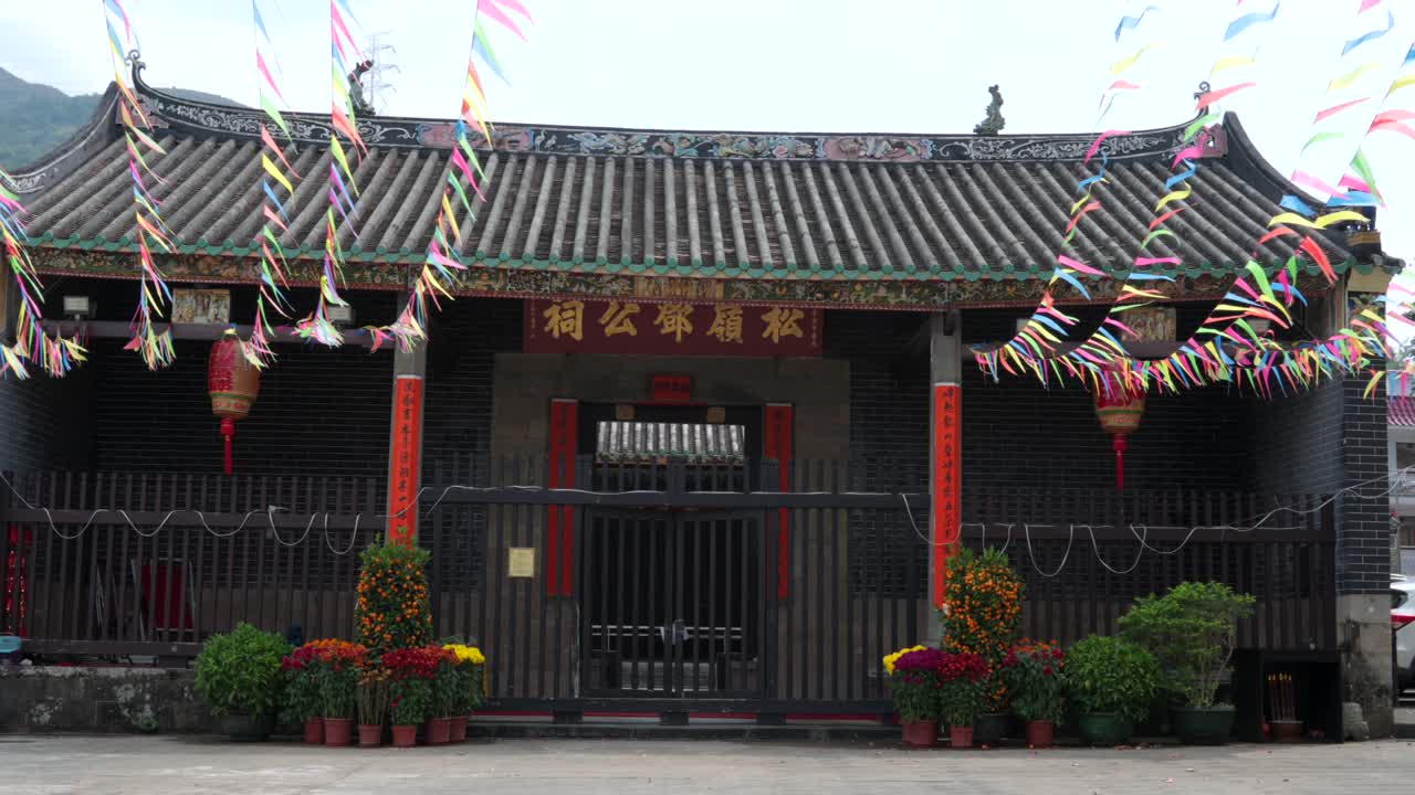 Ornate Chinese gate with swaying lanterns, floral pots, and festive banners under tiled roof.