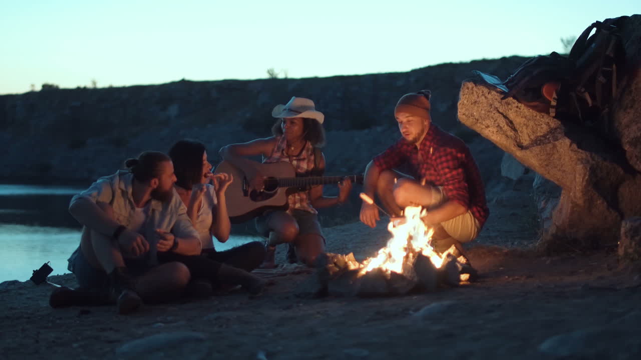 Friends camping by the lake with a campfire, singing and playing guitar