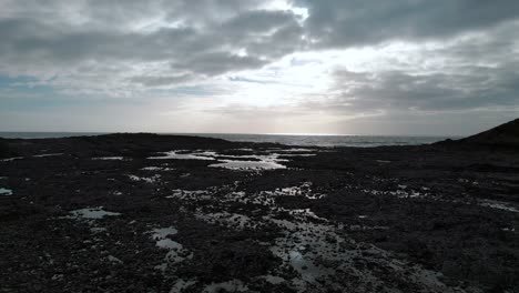 AERIAL:-Low-dolly-towards-sea-over-textured-rockpools-with-sunrays-through-clouds,-Port-Eynon-Gower,-4k-Drone