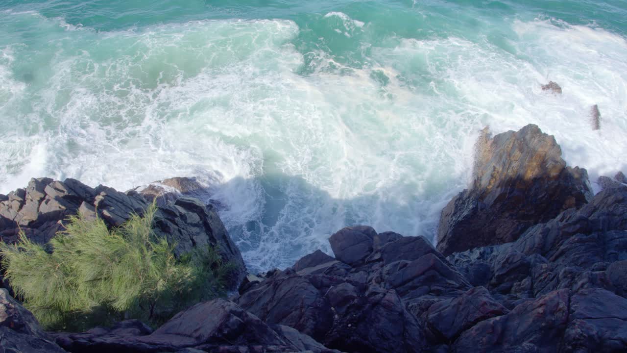 una vista desde un acantilado rocoso con vistas a las olas turquesas que se estrellan contra las rocas oscuras. un pequeño parche de vegetación se aferra al acantilado.