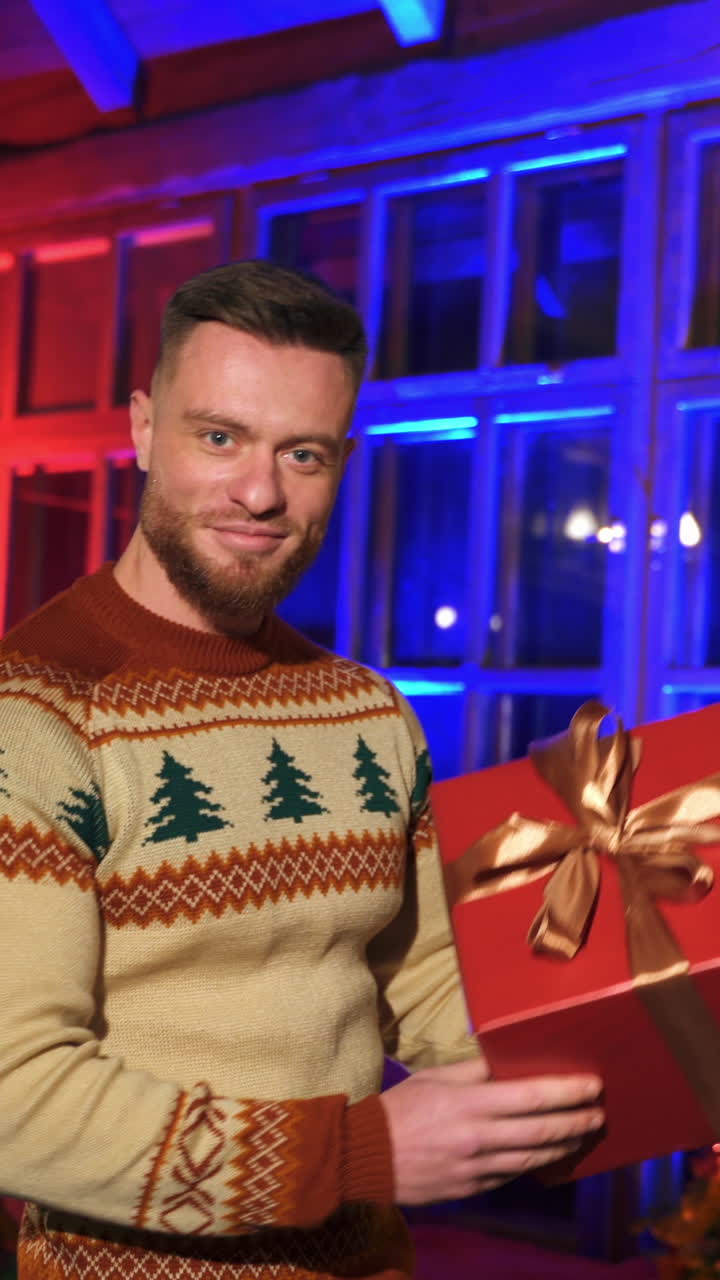 Smiling man offering new year present. Portrait of a handsome man in knitted Christmas sweater holding a box with gift on Christmas decorated room background. Vertical video