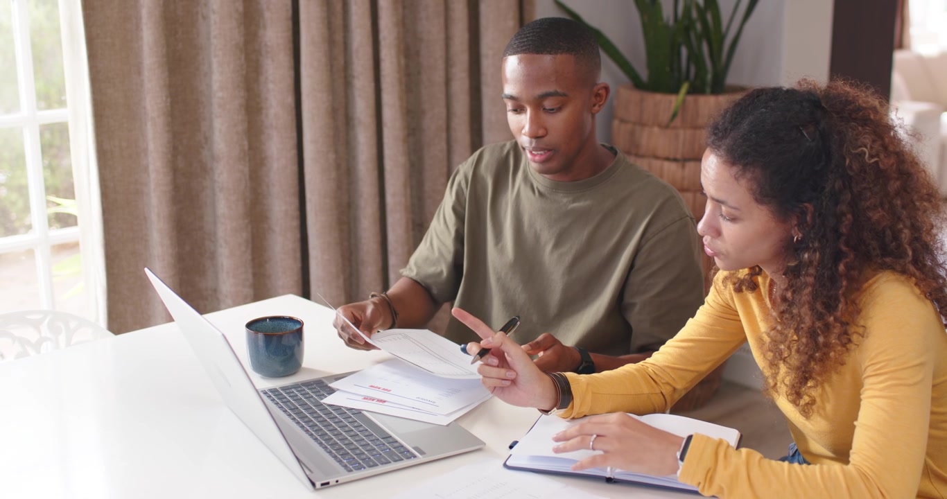 Diverse couple reviewing laptop and papers after opening mail at home on white table taking notes