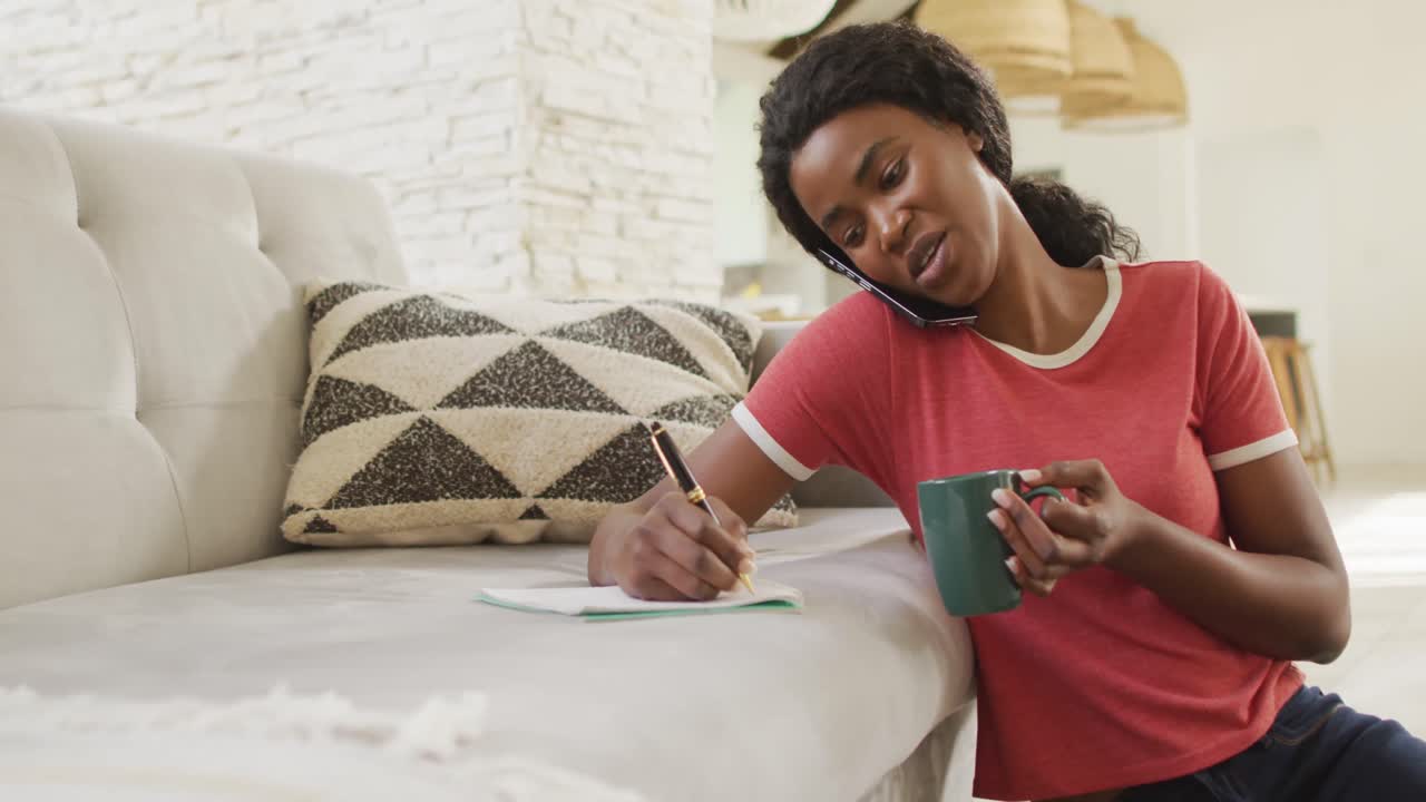 Video di donna afroamericana felice seduta sul pavimento, che usa lo smartphone e prende appunti