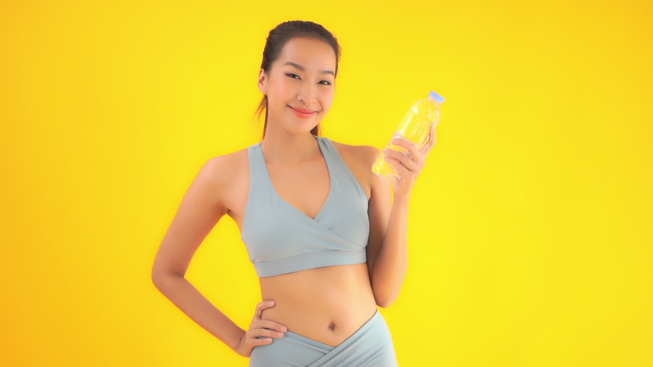 Beautiful happy asian woman in sport suit showing bottle of water to camera, full frame slow motion. Isolated on yellow background