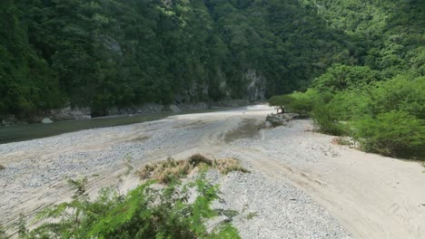 Aerial-forward-at-low-altitude-over-riverbank-at-Muchas-Aguas-with-people-in-background,-Dominican-Republic