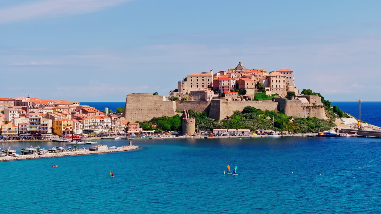 Aerial drone shot over the coastal town of Calvi in Corsica. View of the citadel fortress overlooking the city. Summer holidays destination. Bright blue sky, vibrant turquoise sea