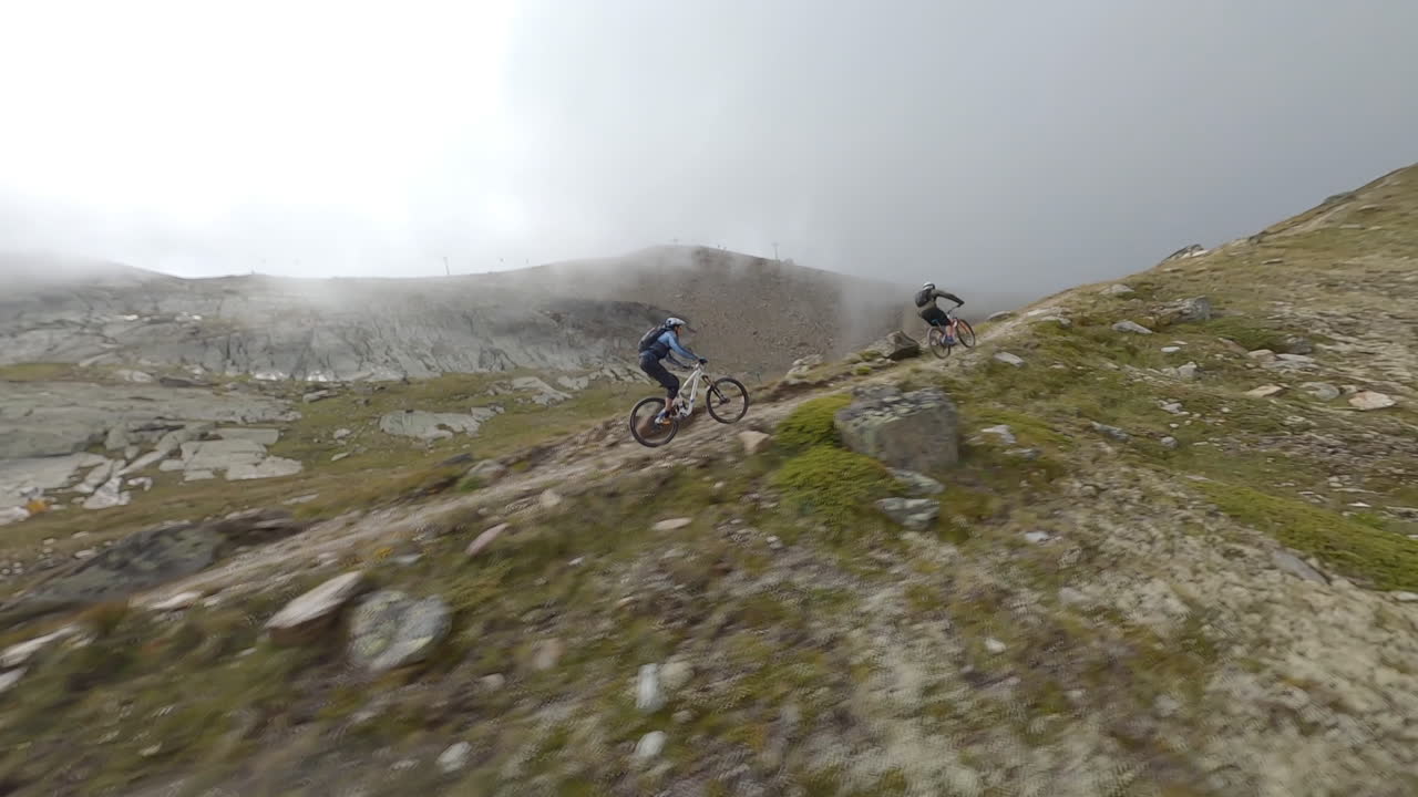 FPV drone follows two mountain bikers descending steep, exposed alpine trails above Saas-Grund and Saas-Fee on a clear bluebird summer day, showcasing epic terrain and speed