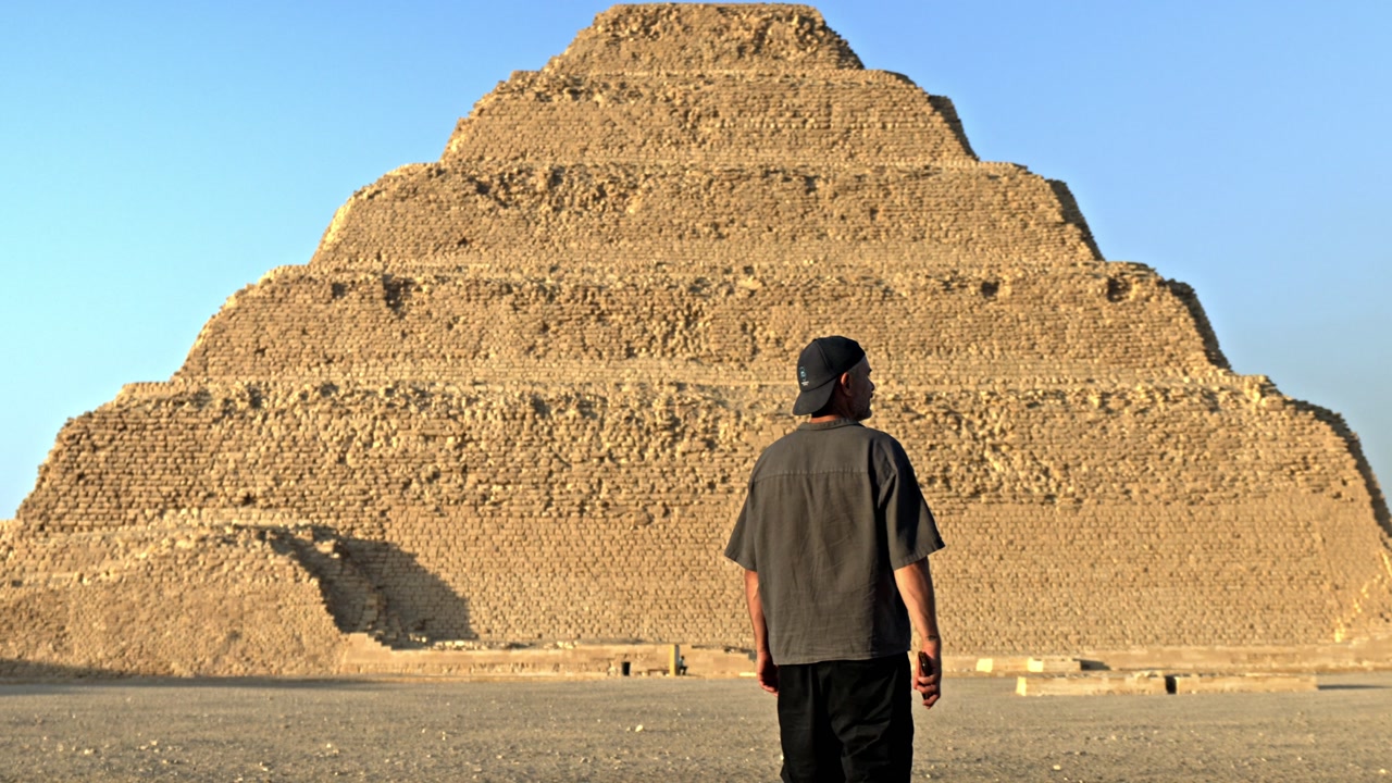 Man walking across the desert plateau of Saqqara as the sun sets, casting a warm, golden glow over the ancient landscape. In the background stands the iconic Step Pyramid of Djoser.