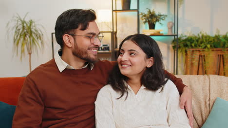 loving happy smiling young indian couple spending leisure time sitting arm around relaxing on sofa