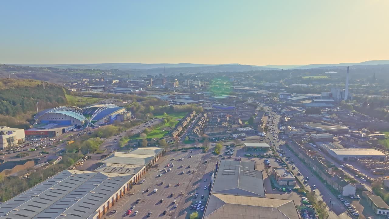 Wide drone shot captures Huddersfield cityscape including John Smith's Stadium, surrounding residential, industrial zones, and the distant Pennine hills under soft golden afternoon light, aerial view