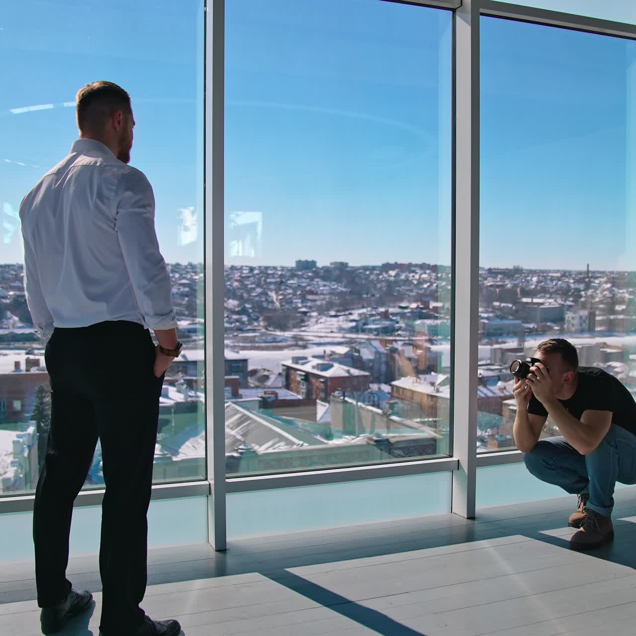 Professional photographer shooting businessman. Handsome man in black trousers and white shirt posing on camera while standing indoors near windows