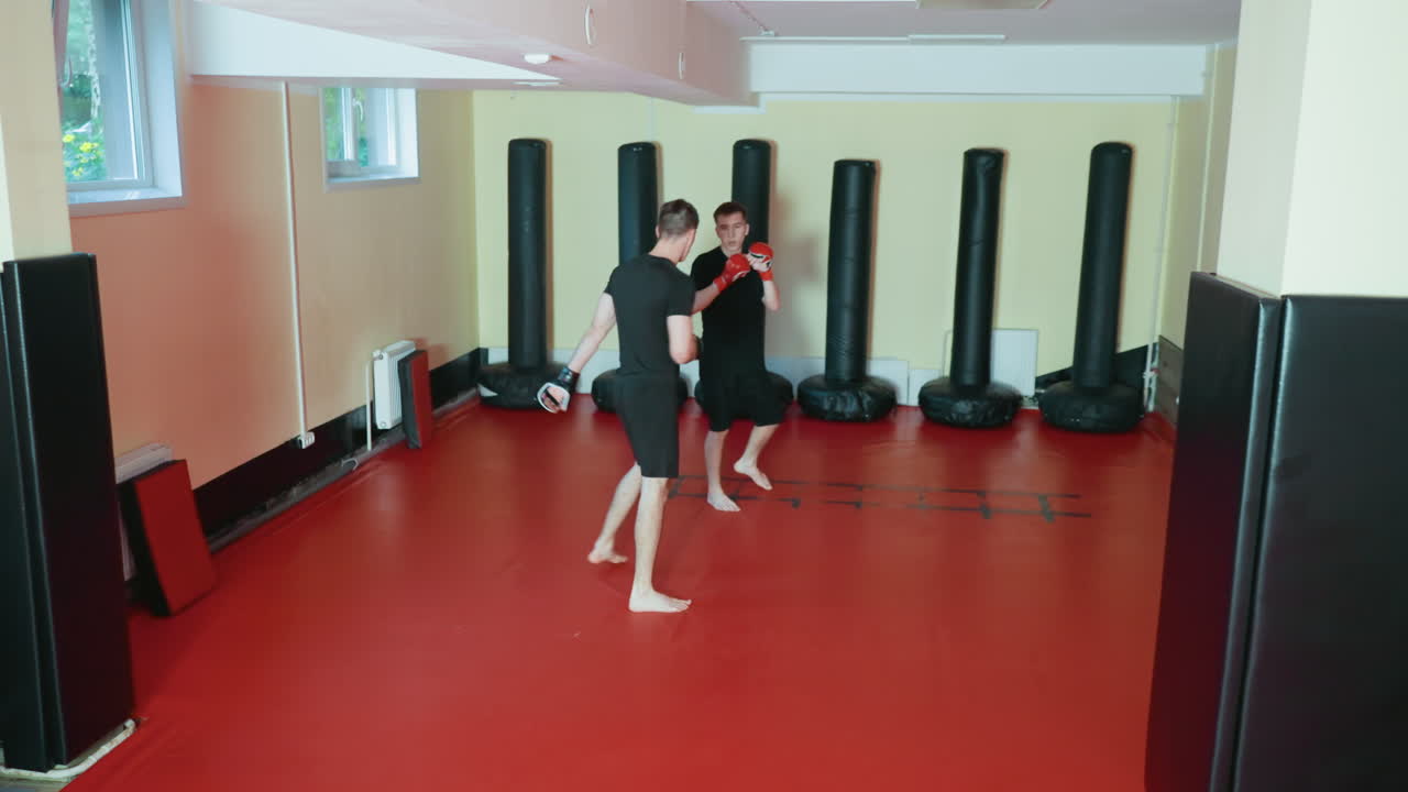 레슬링 선수들이 무술 체육관의 빨간 매트 위에서 맨발로 스파링을 하고 있어요. 한 선수가 하이킥을 날리는 동안 상대 선수는 글러브로 막고 있어요. 샌드백과 패딩 처리된 벽으로 둘러싸인 공간에서 집중하며 격투 기술을 연습하고 있네요.