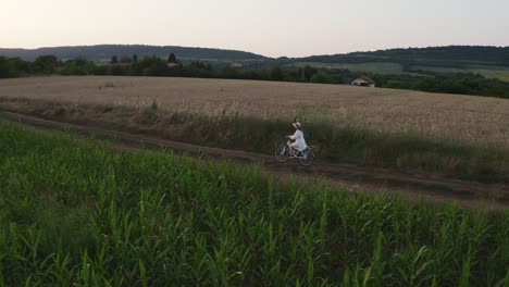 Ein-Einsames-Mädchen-In-Weißem-Kleid-Fährt-Zur-Goldenen-Stunde-Mit-Dem-Fahrrad-Durch-Ländliches-Ackerland