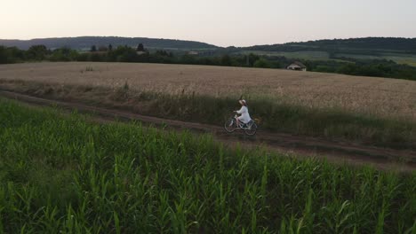 lone girl in white dress rides bike through rural farmland at golden hour