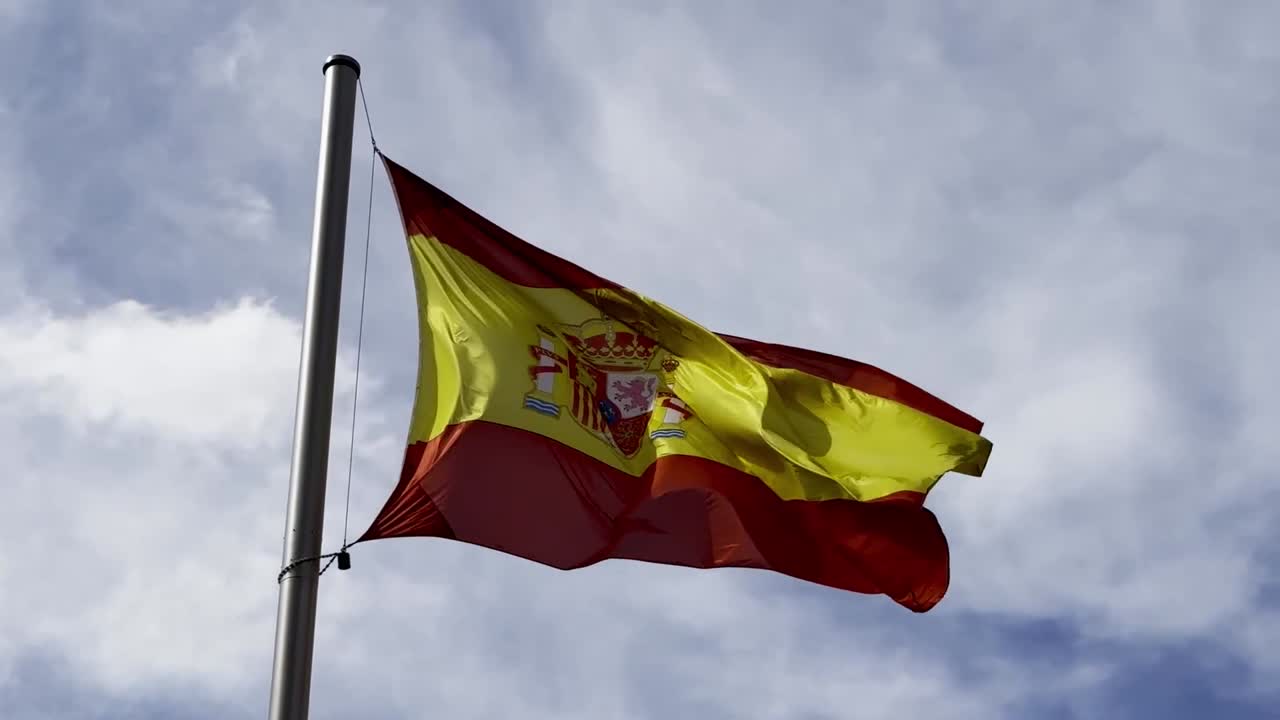 Inquadratura dal basso di una bandiera nazionale che sventola al vento su un'alto pennone con un cielo nuvoloso e drammatico sullo sfondo, a simboleggiare identità e orgoglio in uno spazio pubblico circondato da punti di riferimento storici e culturali