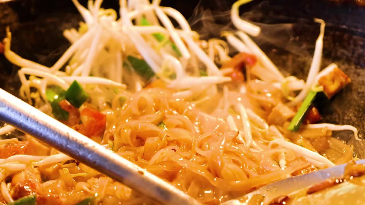 Close-up of noodles and bean sprouts being stir-fried in a hot wok with a metal spatula.