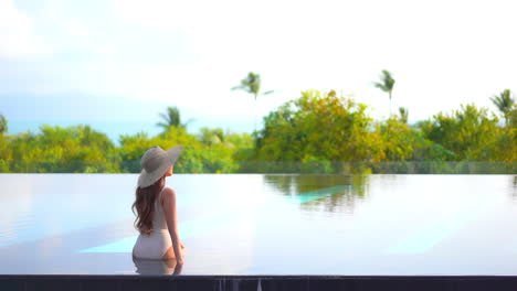vista trasera de mujer sexy en traje de baño y sombrero en piscina infinita con hermosa vista de isla tropical y horizonte oceánico