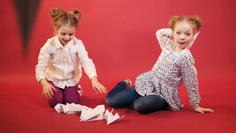 two little girls are playing and throwing origami in the form of planes on the background of red studio