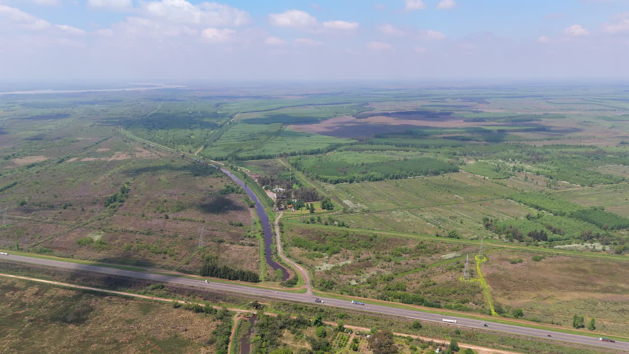 High altitude, wide-angle aerial clip going over Delta del Paraná, highway and vast extension, with clouds and sunny patches, and distant horizon.