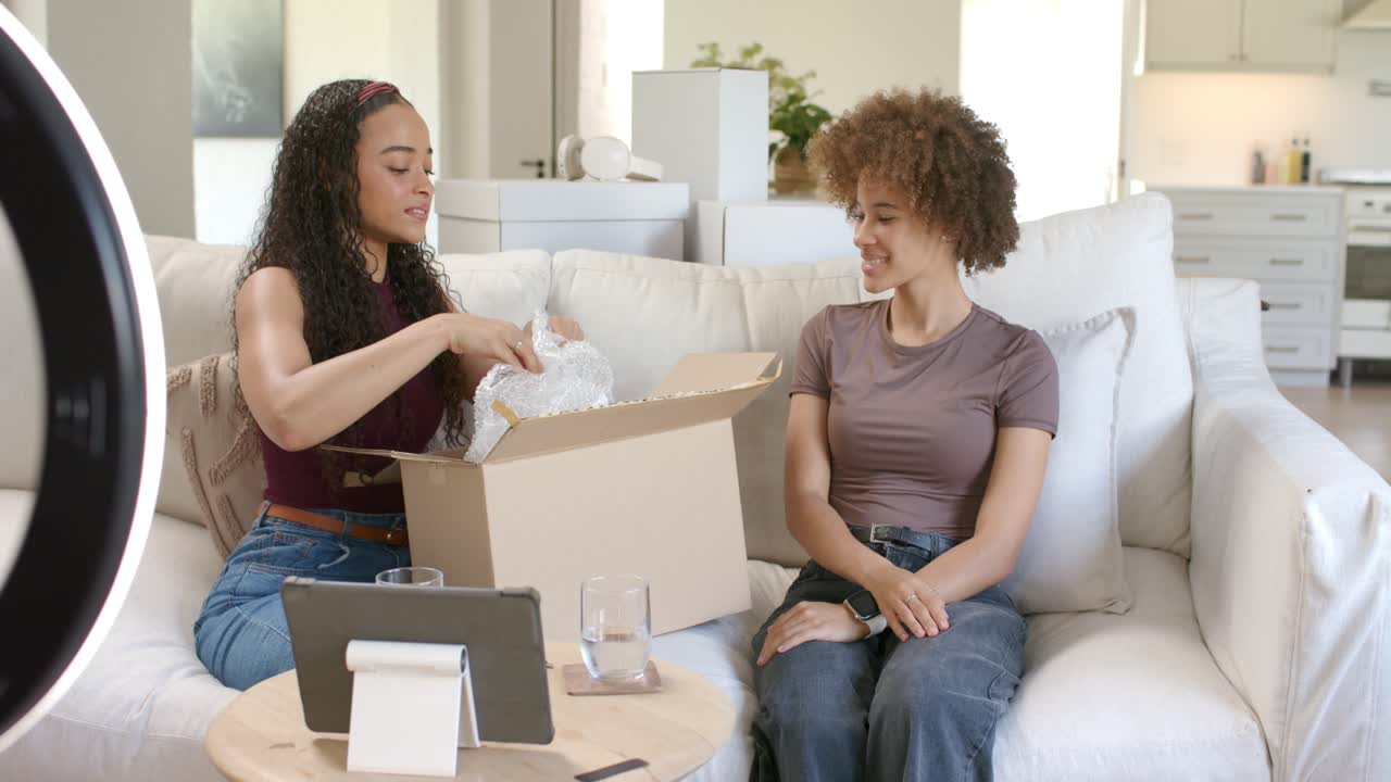 Des femmes afro-américaines déballant un nouveau colis sur un canapé, déchirant le papier bulle pour révéler un appareil destiné à un vlog.
