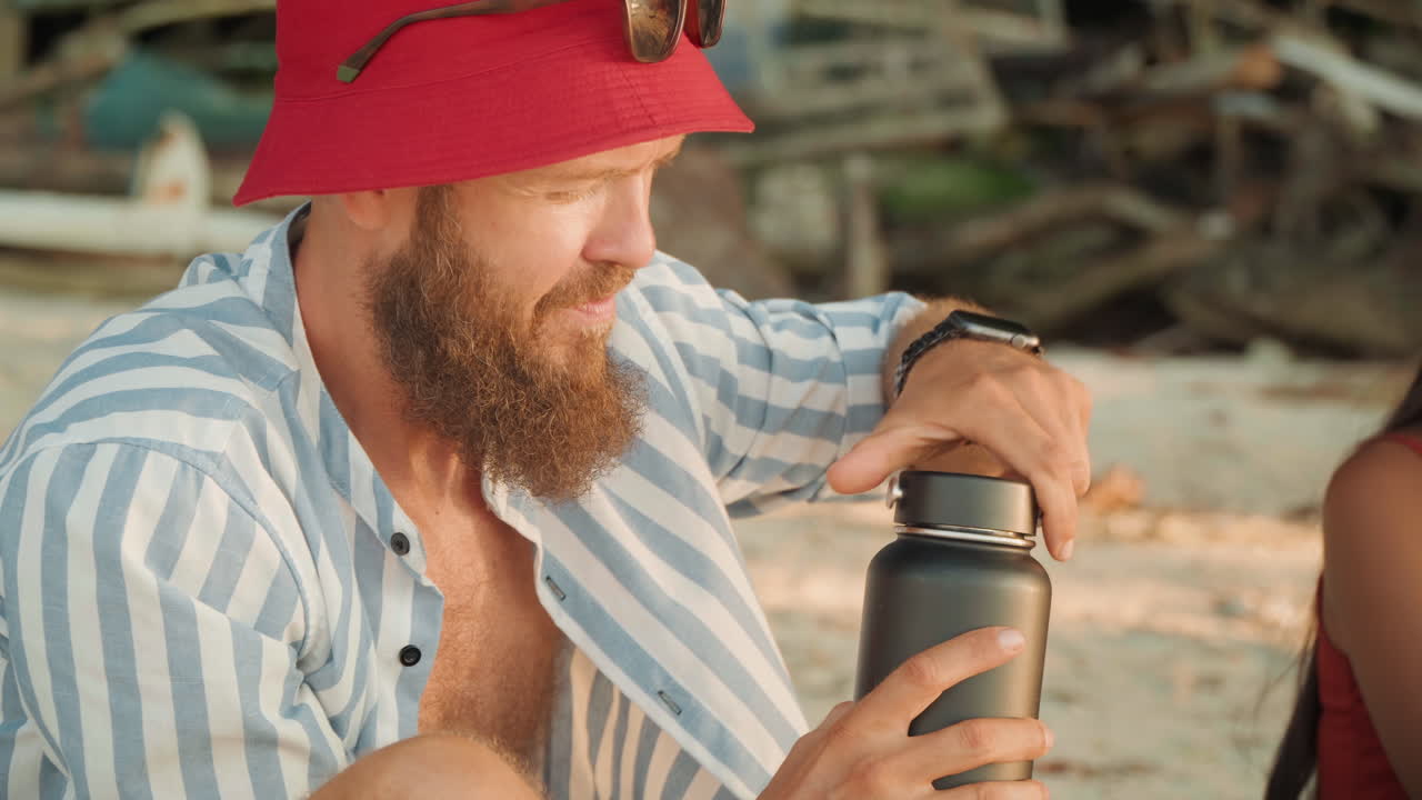 Uomo barbuto che apre un thermos in spiaggia, cappello da pescatore rosso, camicia a righe, sorride mentre svita il tappo della bottiglia e versa da bere, atmosfera da picnic casual, sabbia sotto i piedi, conversazione amichevole, luce del tramonto
