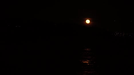 full moon reflection in small waves on water surrounded by deep black background, small city lights in the distance