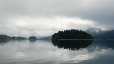 Isla-Boscosa-En-Medio-De-Un-Lago-Con-Reflejos,-Toma-En-Movimiento-Desde-Un-Barco,-Día-De-Mal-Humor,-Isla-De-Vancouver