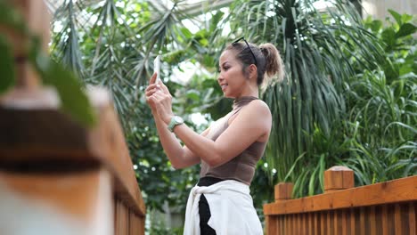 mujer tomando una selfie con su teléfono inteligente en un exuberante jardín interior, rodeada de plantas verdes