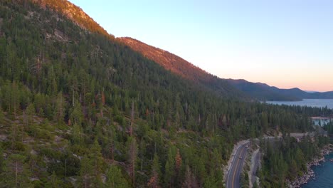 Coches-Conduciendo-Por-La-Carretera-De-Montaña-A-Través-Del-Bosque-De-Pinos-De-Jeffrey-Y-Las-Aguas-Azules-Del-Lago-Tahoe-Al-Atardecer-En-California---Toma-Aérea-De-Drones