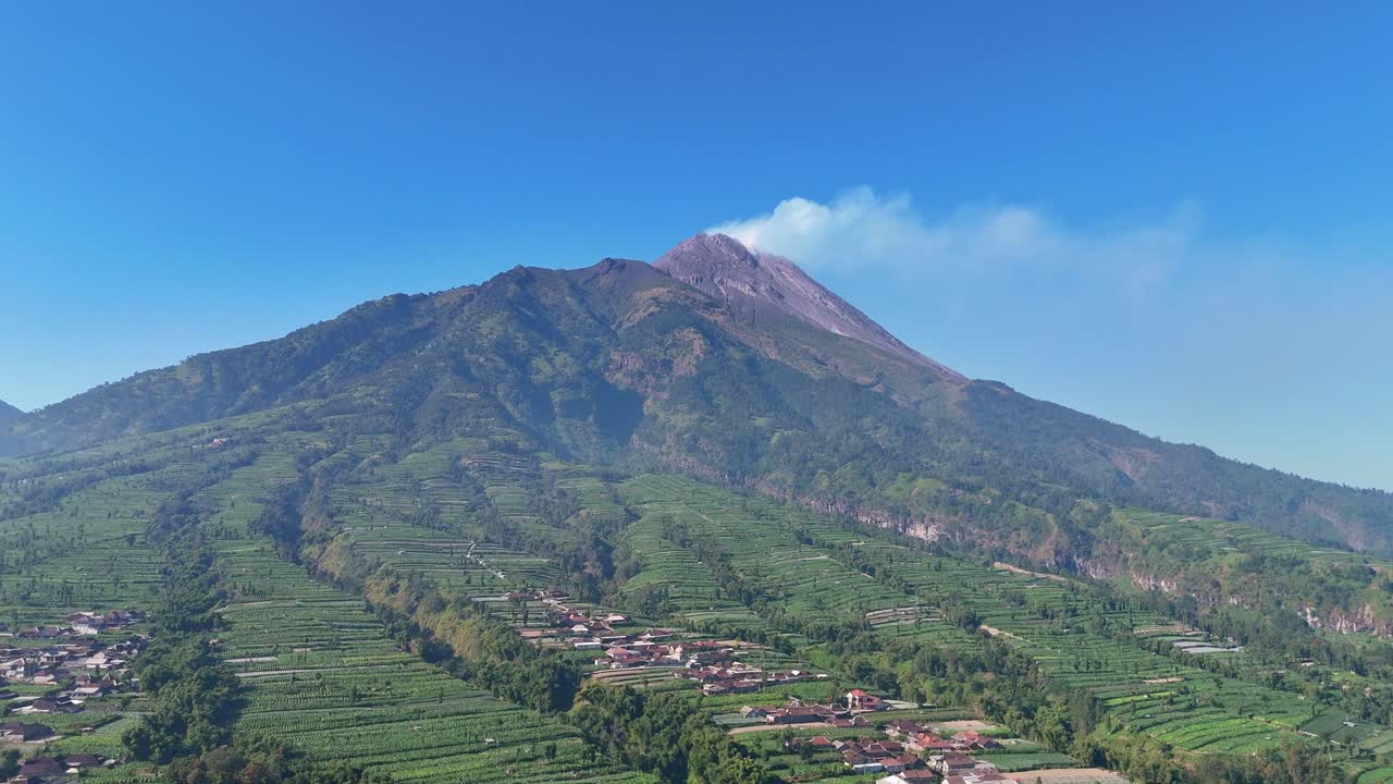 Aerial scenery of majestic volcano emitting white smoke above rural farmland under blue sky. Mount Merapi Volcano, Indonesia