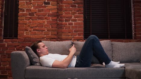 the young serious looking man is lying on the couch in the living room and is writing a message with his smartphone, shot from the side