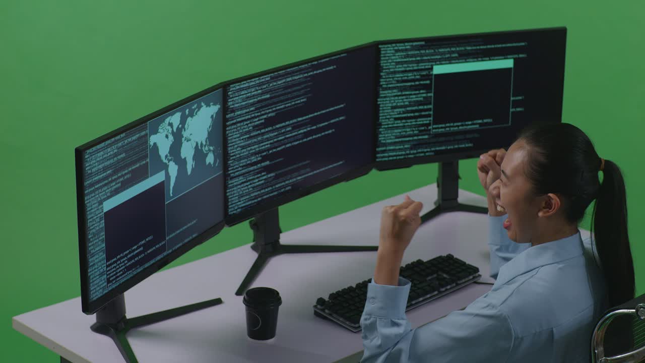 vista lateral de una desarrolladora asiática feliz celebrando mientras escribe código con múltiples pantallas de computadora en el estudio de pantalla verde